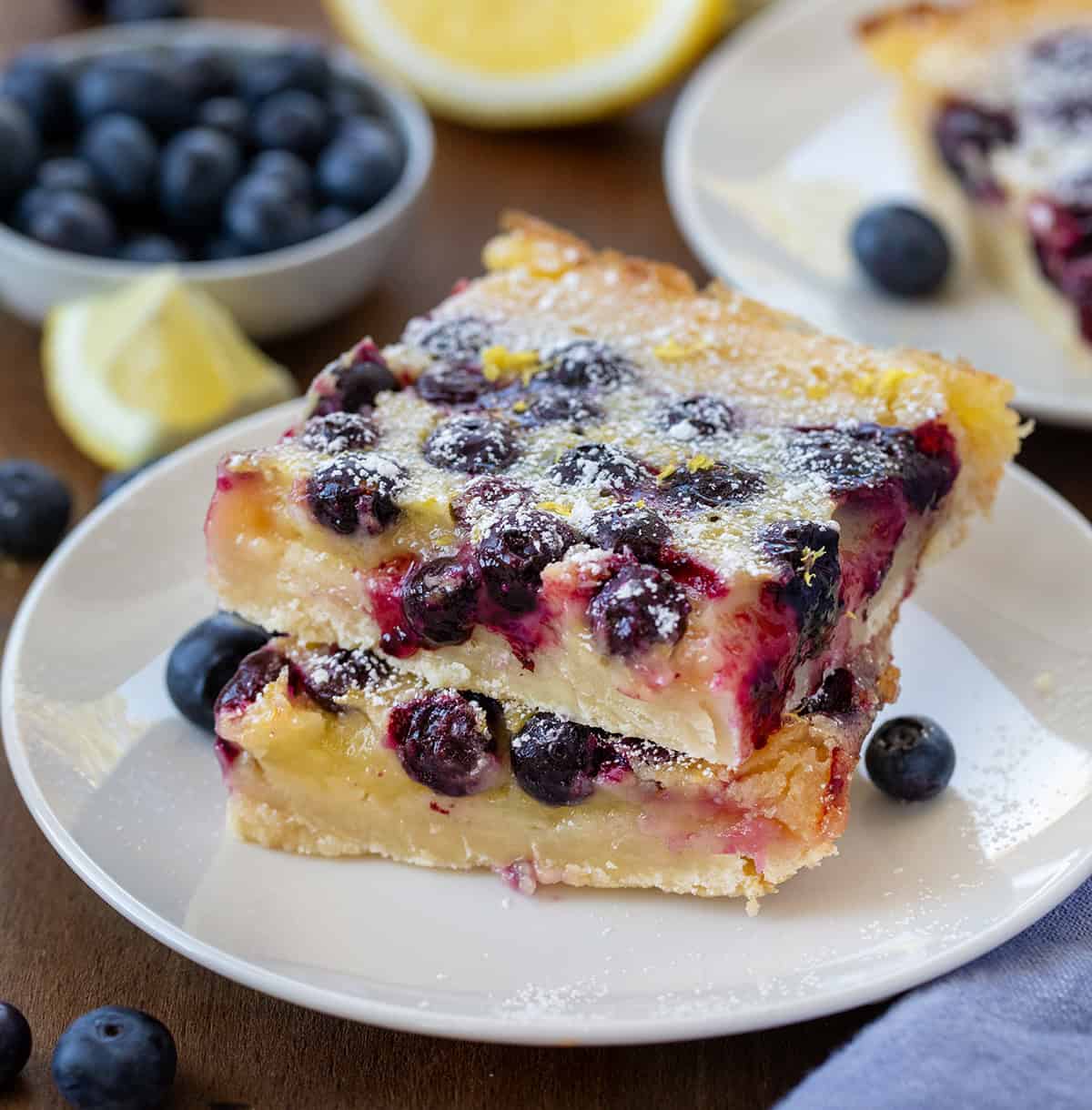 Lemon Blueberry Bars stacked on a white plate on a wooden table surrounded by other bars and fresh fruit.