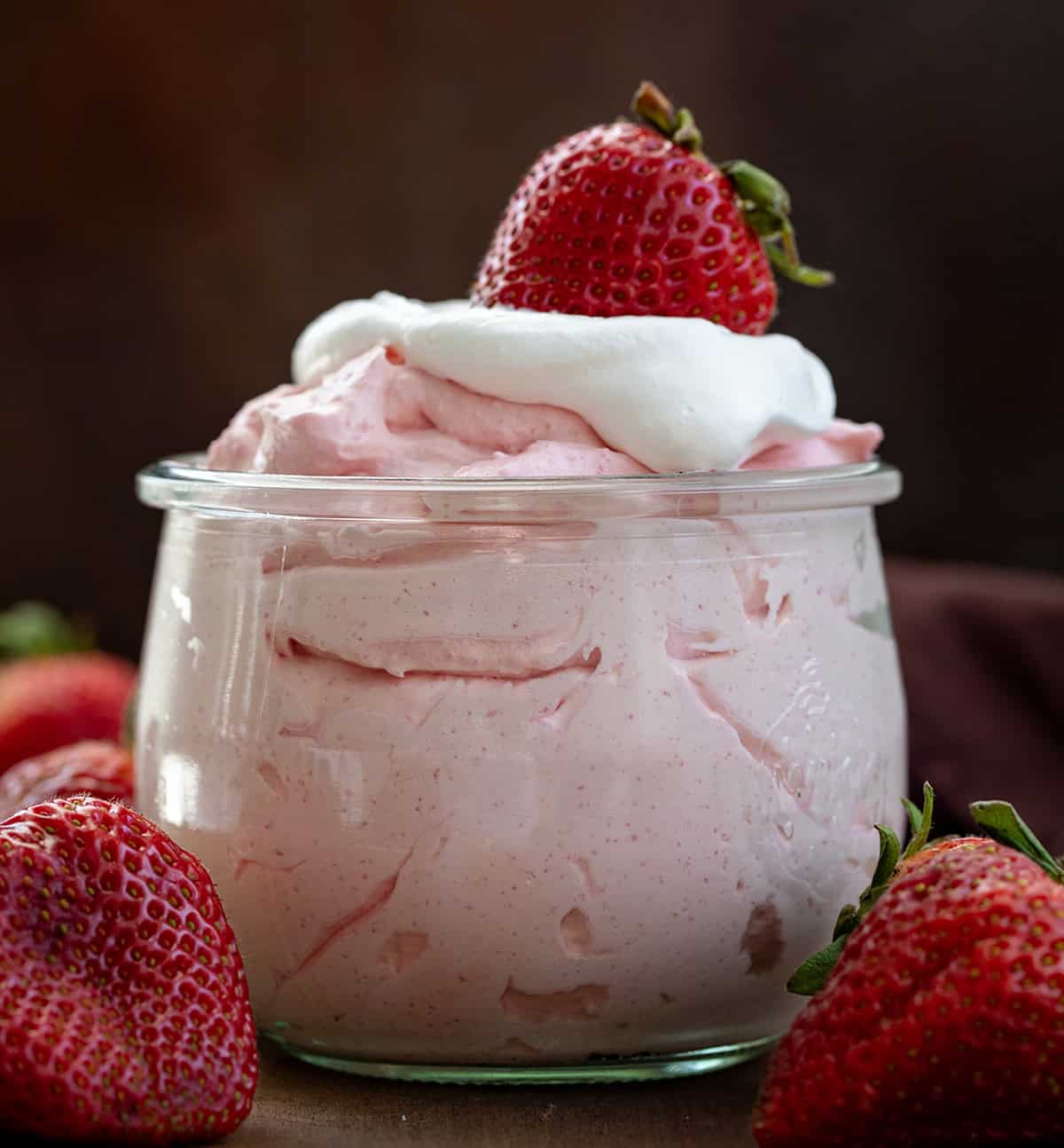 Jar of Strawberry Cheesecake Mousse with whipped cream and a fresh strawberry on top on a wooden table. 