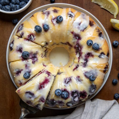 Lemon Blueberry Pound Cake on a wooden table from overhead with pieces cut and one piece being lifted off of the cake plate.