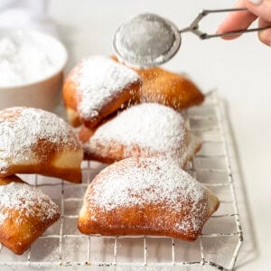 Dusting Beignets with confectioners sugar on a cooling rack on a white counter.