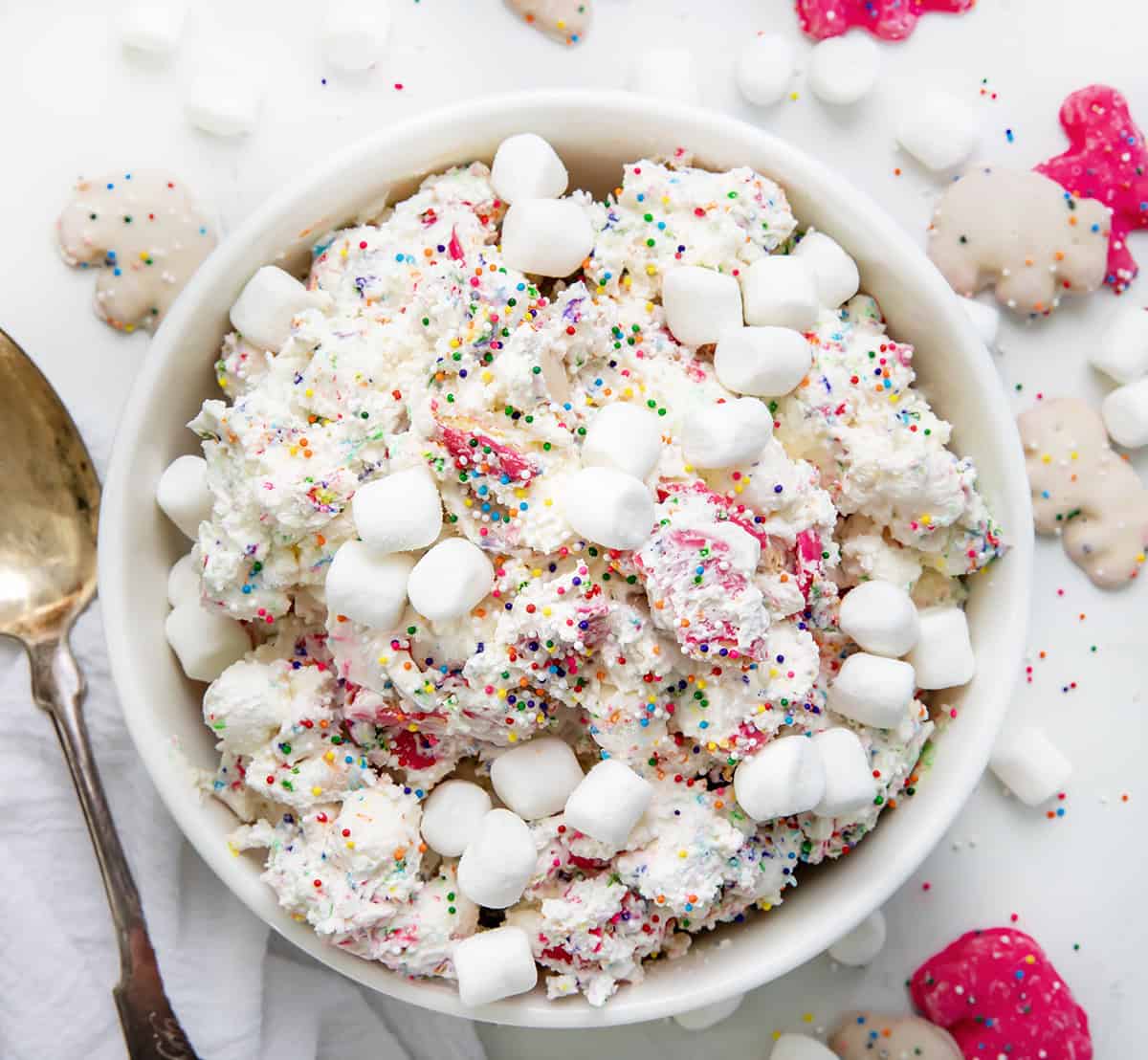 Bowl of Circus Cookie Salad Fluff on a white counter with rainbow non perils and mini marshmallows all around. 
