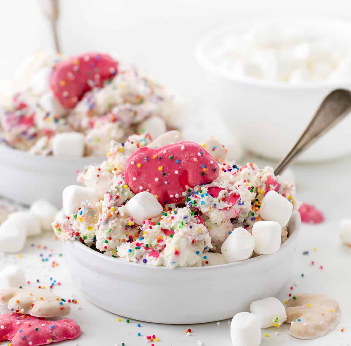 Bowl of Circus Cookie Salad Fluff on a white counter with rainbow non perils and mini marshmallows and all around and spoons.