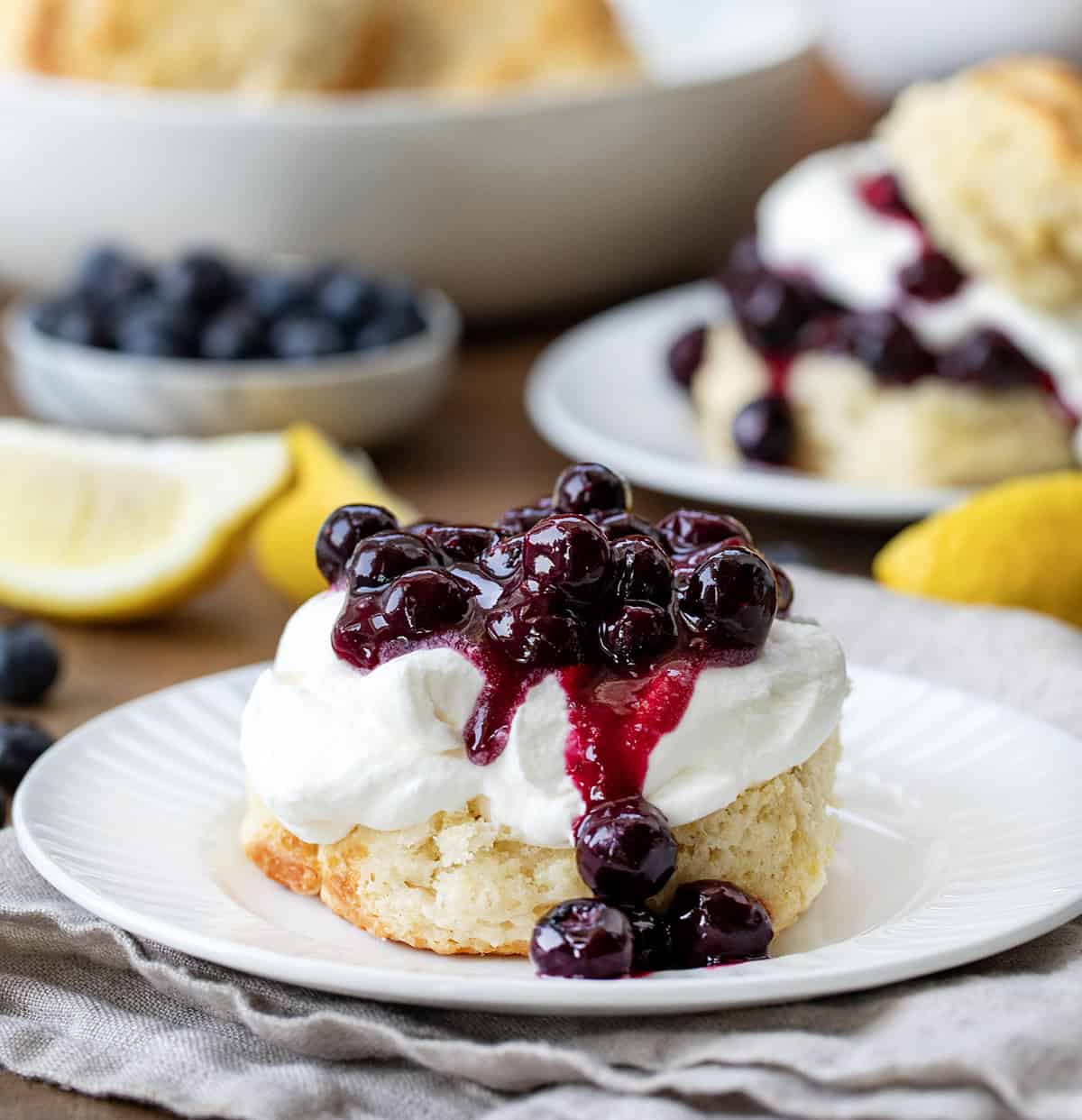 One half of a biscuit topped with whipped cream and blueberry sauce on a white plate on a wooden table with other ingredients surrounding.