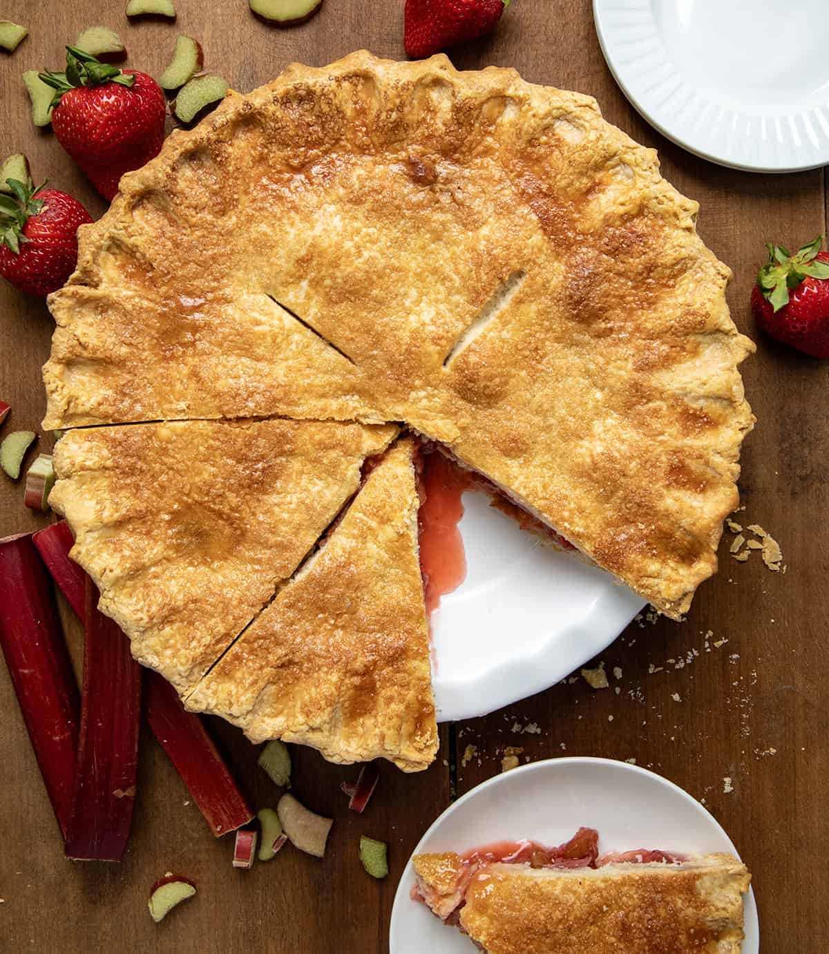 Strawberry Rhubarb Pie on a wooden table with a piece removed and on a plate next to it. 