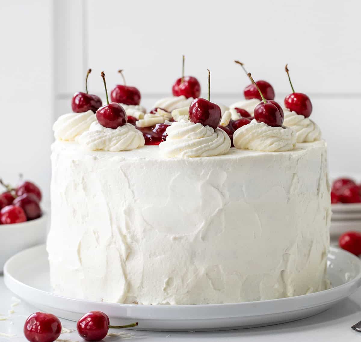 Whole White Forest Cake sitting on a cake plate with fresh cherries all around and white plates in the background.