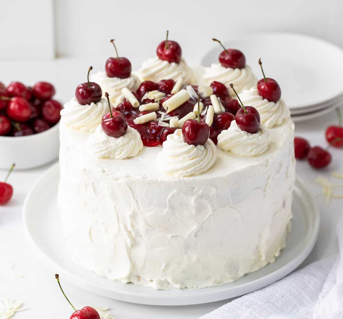 Whole White Forest Cake sitting on a cake plate with fresh cherries all around and white plates in the background. 