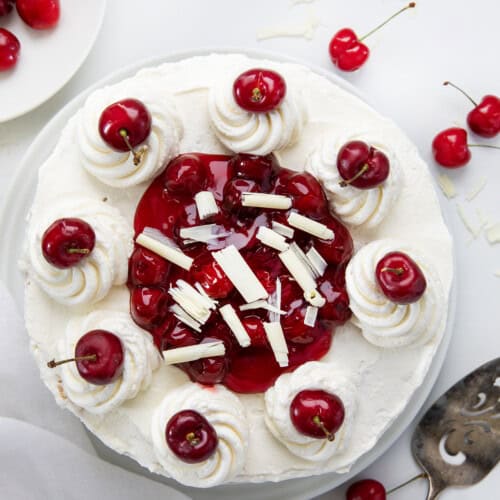 Whole White Forest Cake sitting on a cake plate with fresh cherries all around and white plates in the background from overhead.