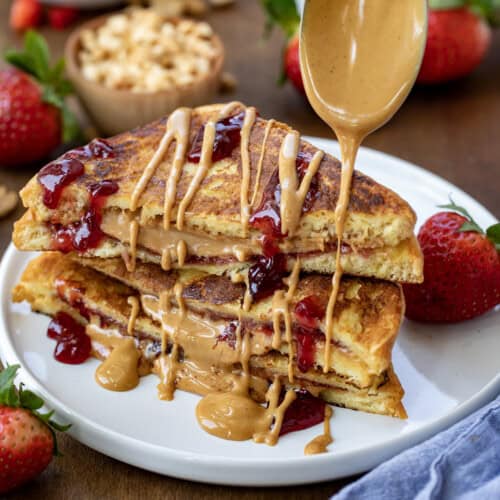 Drizzling peanut butter over Peanut Butter and Jelly French Toast on a plate on a wooden table with fresh strawberries and chopped nuts nearby.