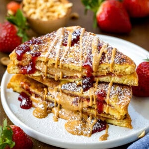 Cut in half and stacked Peanut Butter and Jelly French Toast on a plate that has been drizzled in peanut butter and jelly and dusted with confectioners sugar.