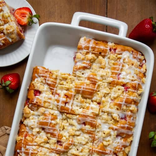 Pan of Strawberry Crumb Cake on a wooden table with one piece removed and next to the pan on a white plate.