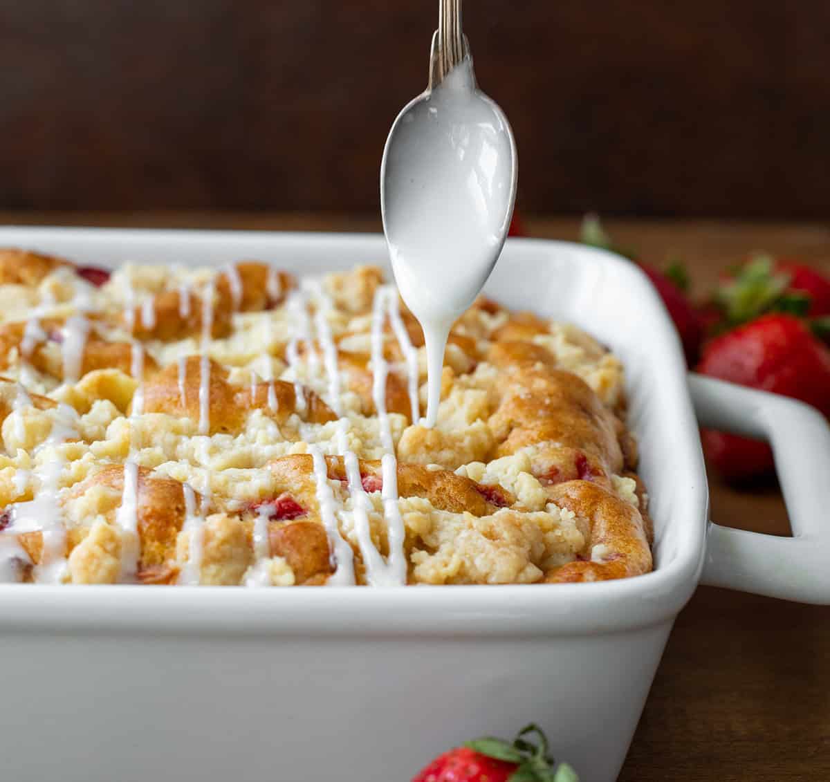 Drizzling glaze over Strawberry Crumb Cake in a white pan on a wooden table.