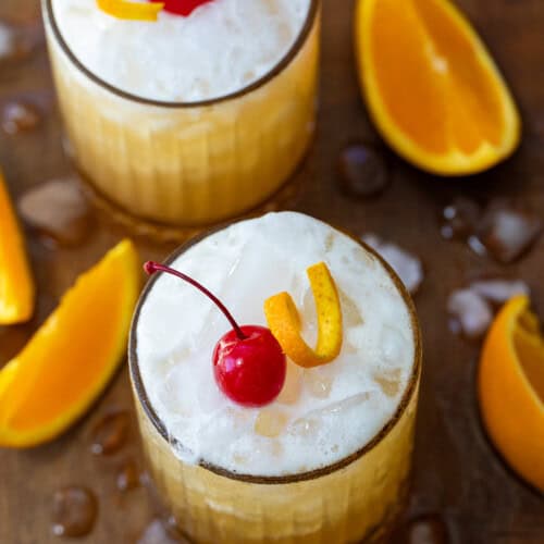 Two Amaretto Sour's on a wooden table with ice and oranges around.