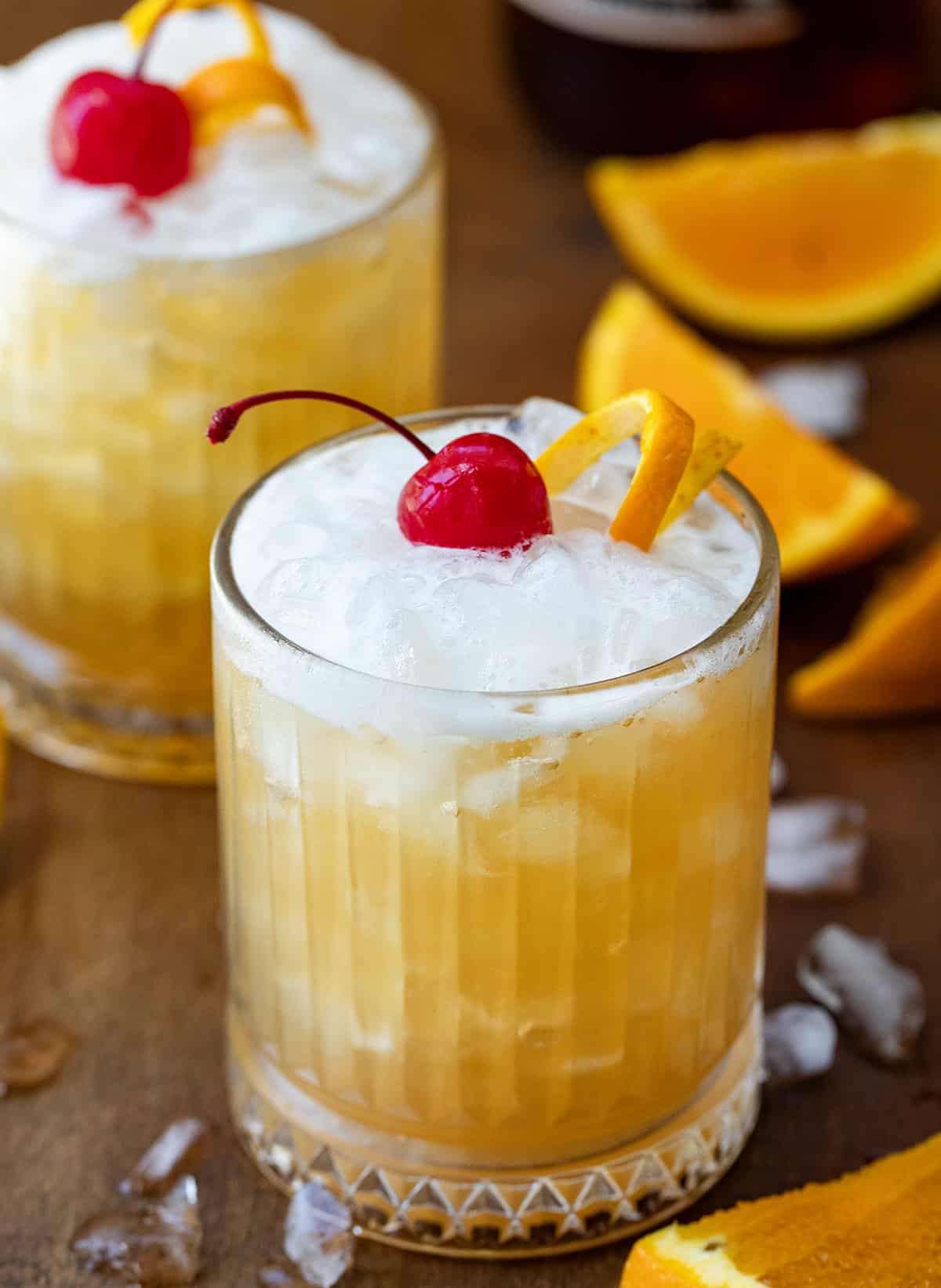 Two Amaretto Sour's on a wooden table with ice and oranges around.