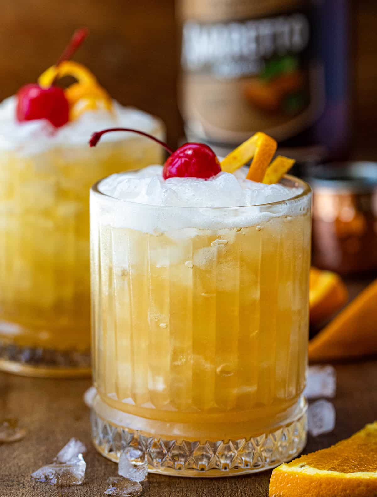 Two Amaretto Sour's on a wooden table with ice and oranges around.