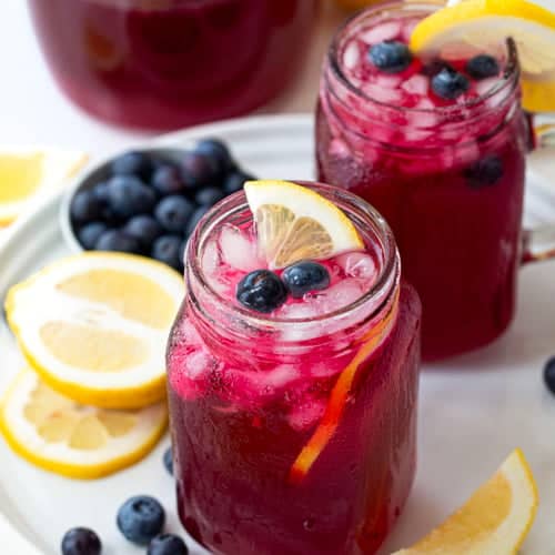 Glasses of Blueberry Lemonade on a white table with fresh lemon and blueberry around.