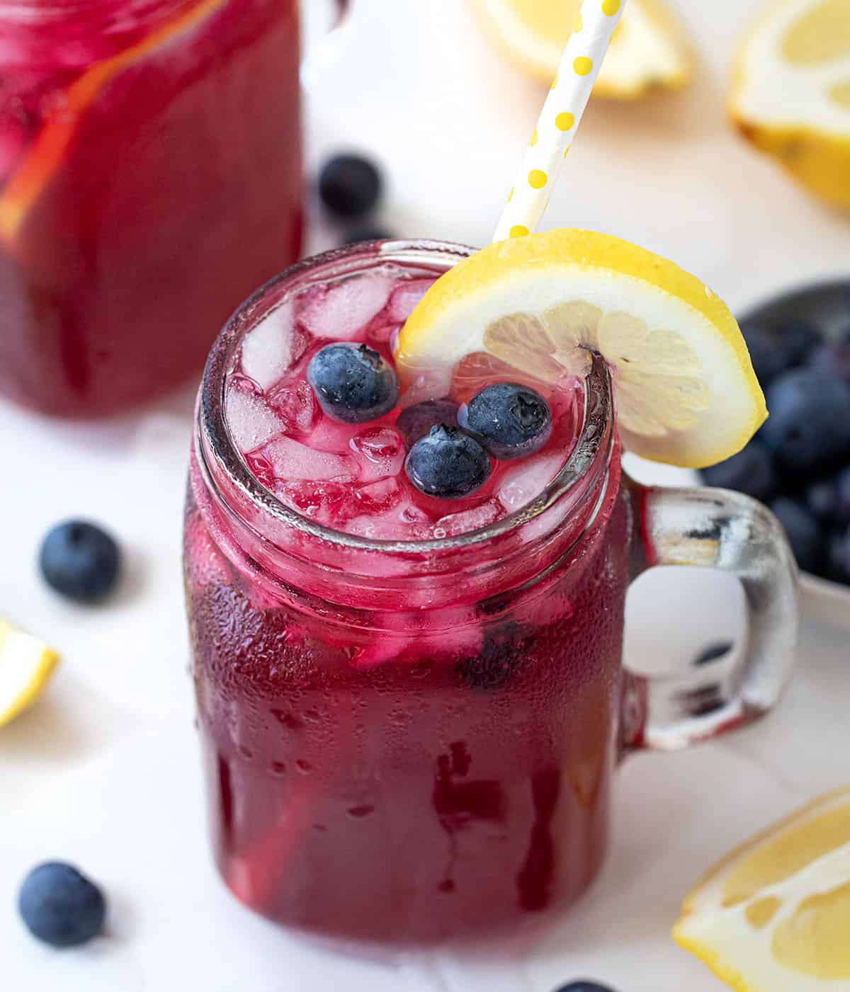 Glasses of Blueberry Lemonade on a white table with fresh blueberries and lemons around.