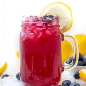 Glass of blueberry lemonade on a white table with a lemon and fresh blueberry garnish.