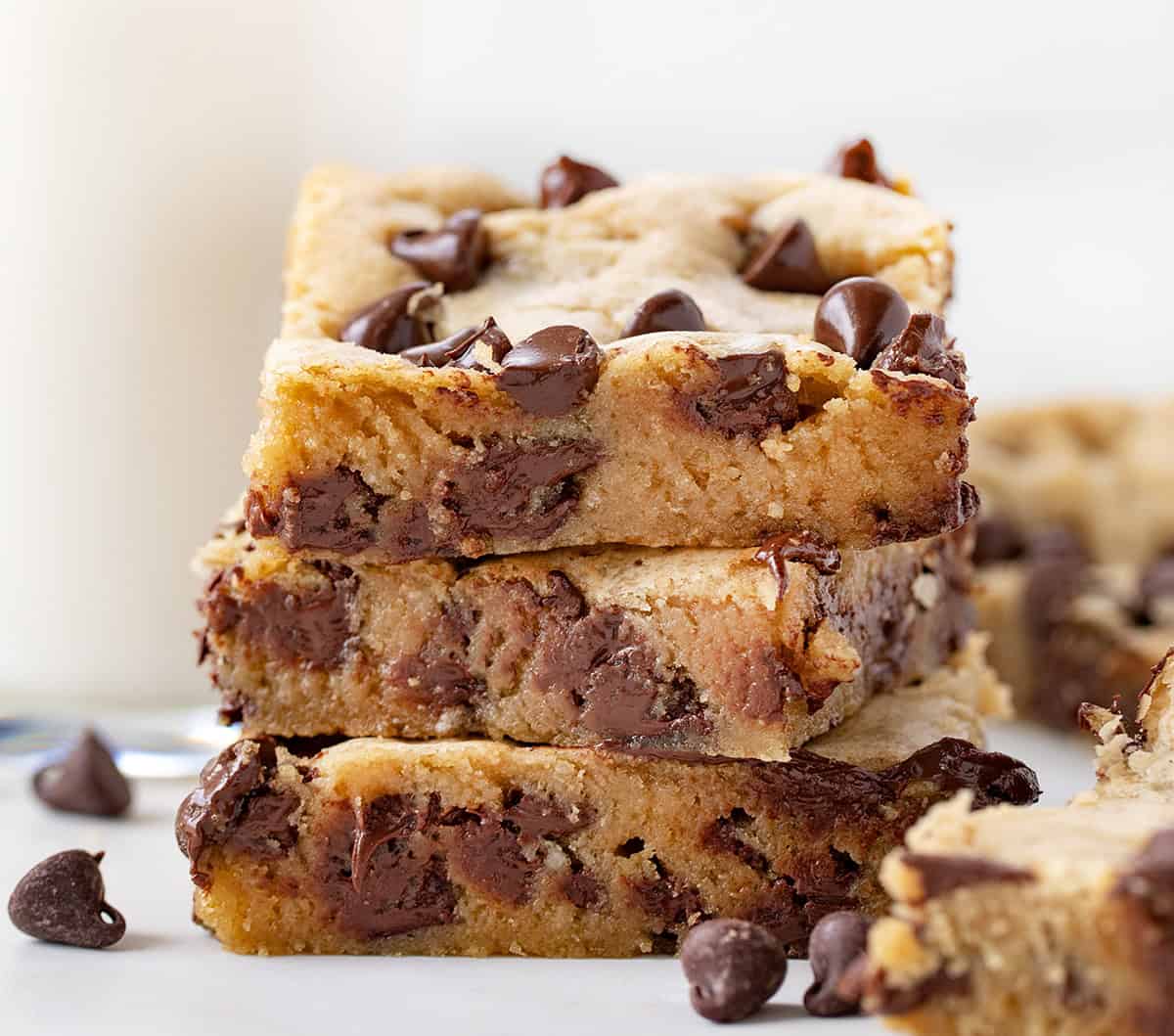 Stack of Browned Butter Chocolate Chip Cookie Bars on a white table with a glass of milk and chocolate chips.