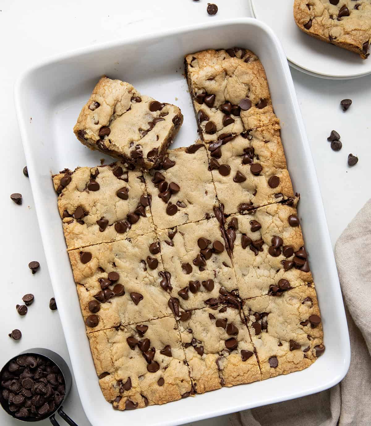Pan of Brown Butter Chocolate Chip Bars on a white table with a napkin and a measuring cup of chocolate chips from overhead. 