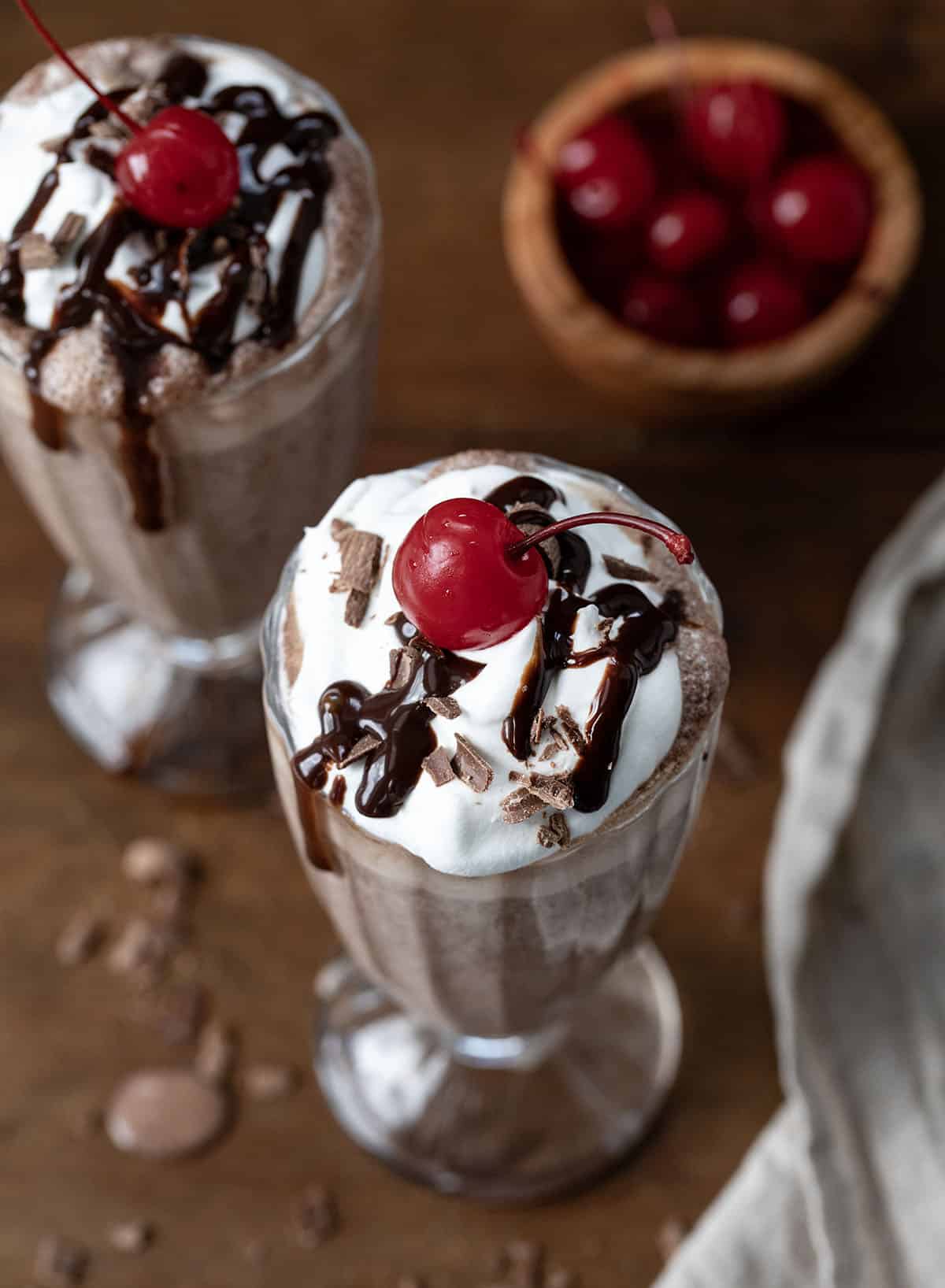 Two Chocolate Milkshakes on a wooden table with hot fudge, whipped cream, and cherry.