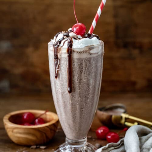 Chocolate MilkShake on a wooden table with whipped cream, hot fudge, cherry, and straw.