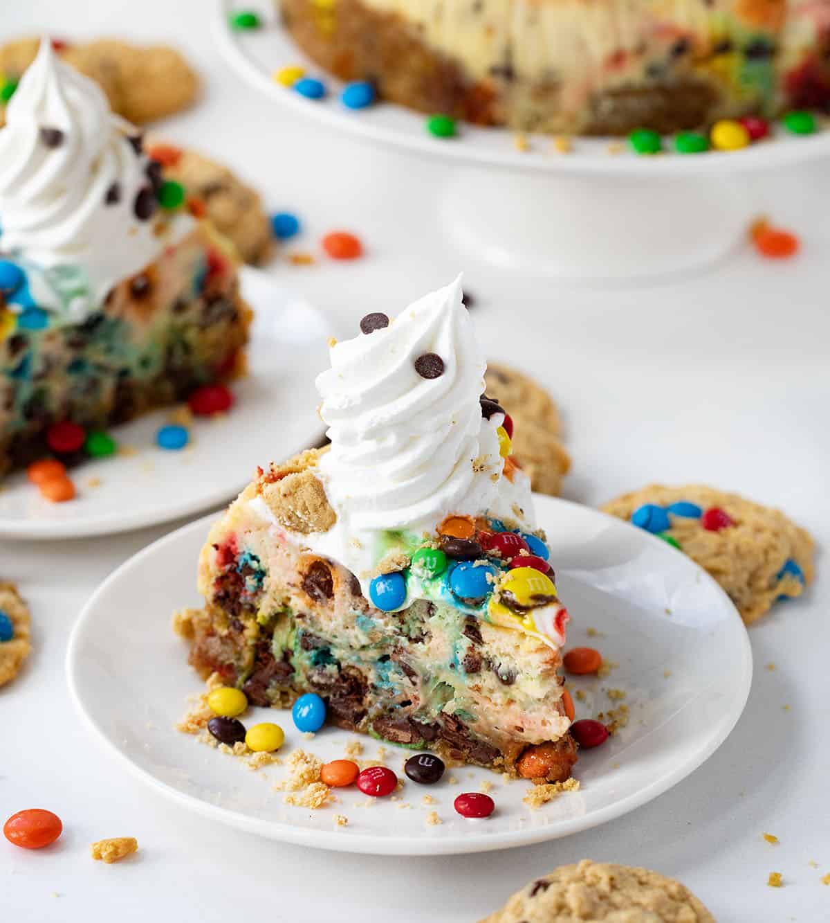 Pieces of Monster Cookie Cheesecake on white plates on a white counter surrounded by cookies.