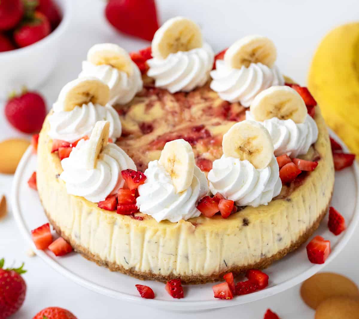 Strawberry Banana Cheesecake on a white cake stand on a white table surrounded by strawberries and banana. 