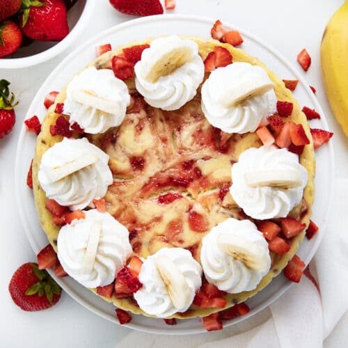 Strawberry Banana Cheesecake on a white cake stand on a white table surrounded by strawberries and banana.