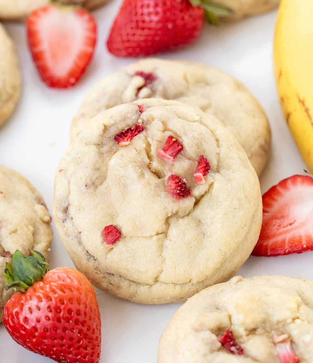 Close up of Strawberry Banana Cheesecake Cookies on a white table surrounded by strawberries and a banana.