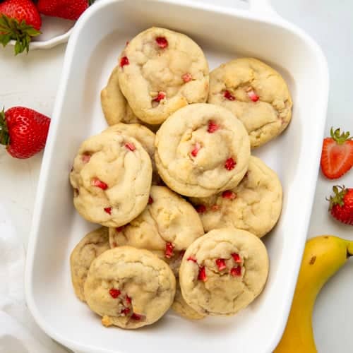 Strawberry Banana Cheesecake Cookies on a white table surrounded by strawberries and a banana.