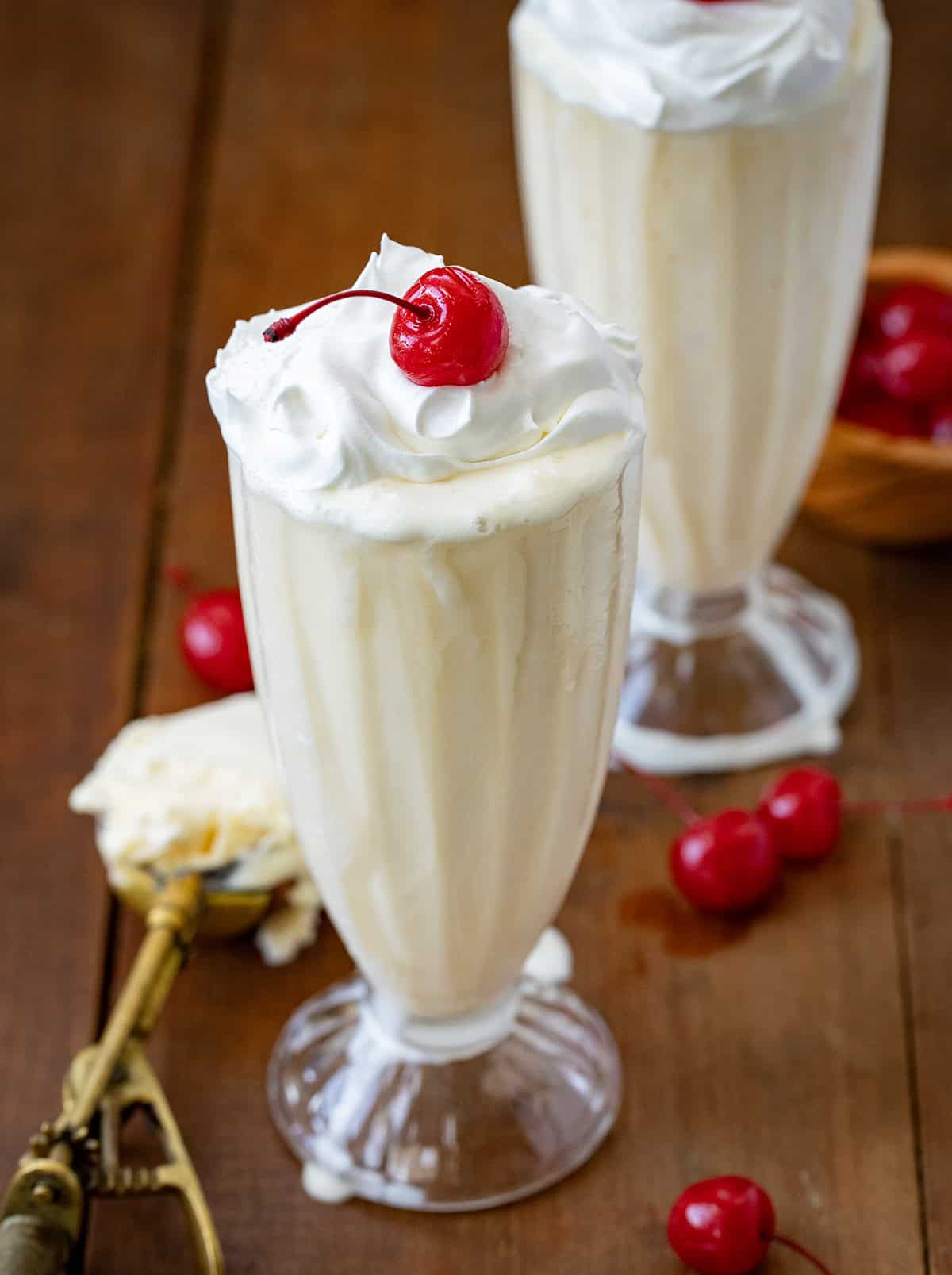 Two Vanilla Milkshakes on a wooden table with whipped cream and cherries.