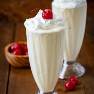 Two Vanilla Milkshakes on a wooden table with whipped cream and cherries.