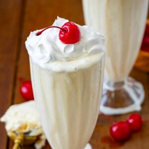 Two Vanilla Milkshakes on a wooden table with whipped cream and cherries.