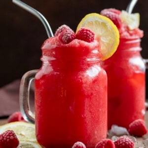 Two Frozen Raspberry Lemonades in mugs with raspberry and lemon garnish on a wooden table.