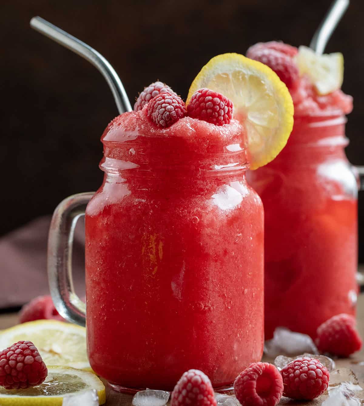 Two Frozen Raspberry Lemonades in mugs with raspberry and lemon garnish on a wooden table. 
