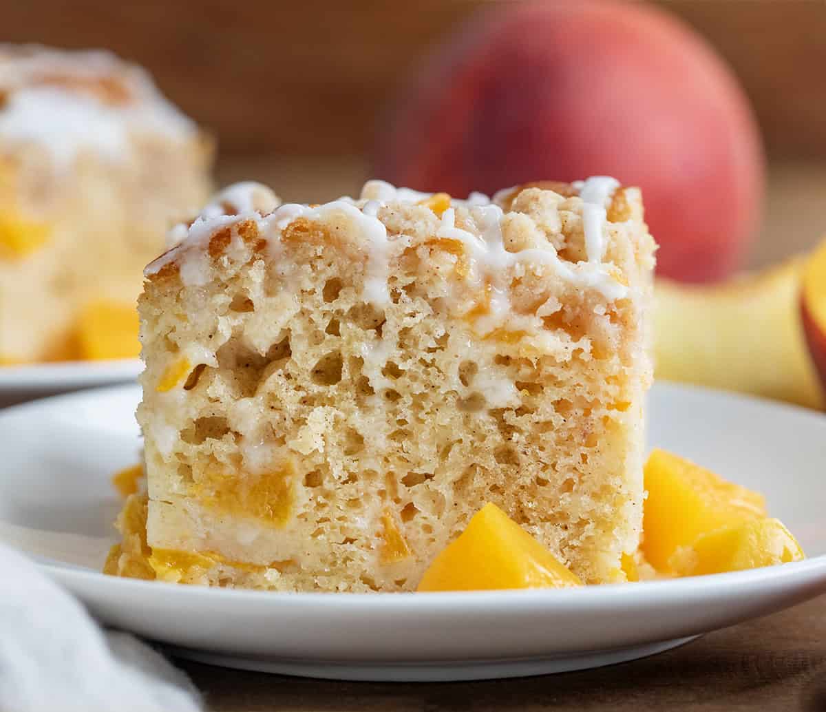 Slice of Peach Crumb Cake on a white plate with peaches in the background.