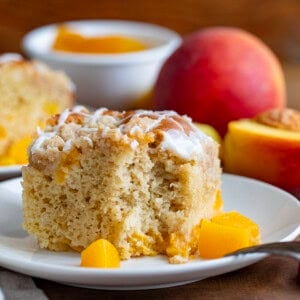 Slice of Peach Crumb Cake on a white plate with peaches in the background.
