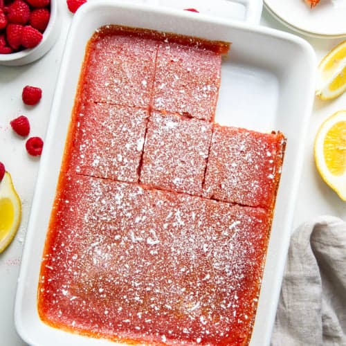 Pan of Raspberry Lemon Bars on a white table with fresh raspberries and lemon wedges from overhead.