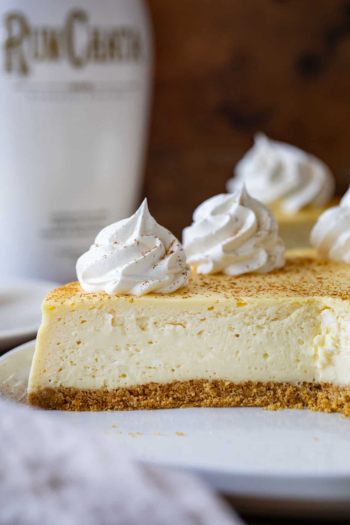 Part of a RumChata Cheesecake on a white cake plate with a bottle of RumChata in the background.