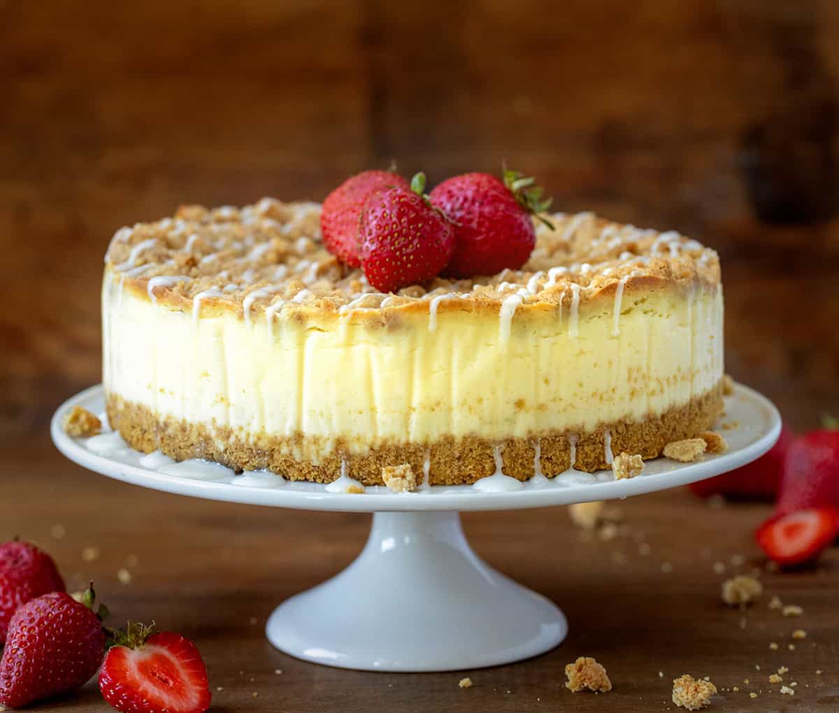 Whole Strawberry Crumble Cheesecake on a cake stand on a wooden table with fresh strawberries. 