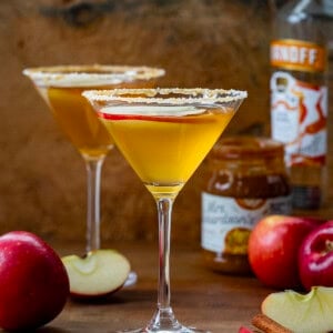 Salted Caramel Apple Martini glasses on a wooden table with caramel, vodka, and apples.