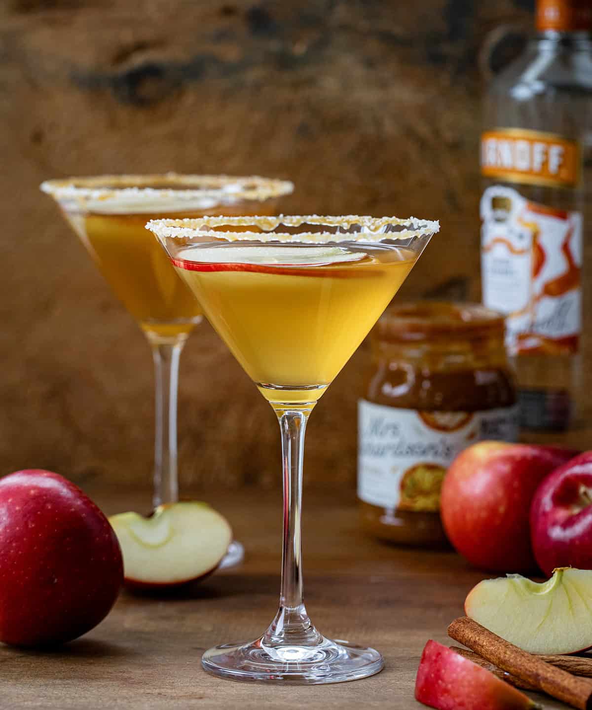 Salted Caramel Apple Martini glasses on a wooden table with caramel, vodka, and apples.