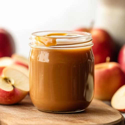 Jar of Apple Cider Caramel Sauce on a wooden table surrounded by apples.