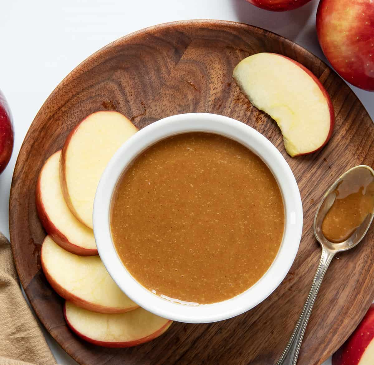 White cup of Apple Cider Caramel Sauce on a wooden plate with fresh apples.