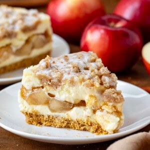 Slice of Apple Crumble Cheesecake on a white plate with another piece in the background with apples.