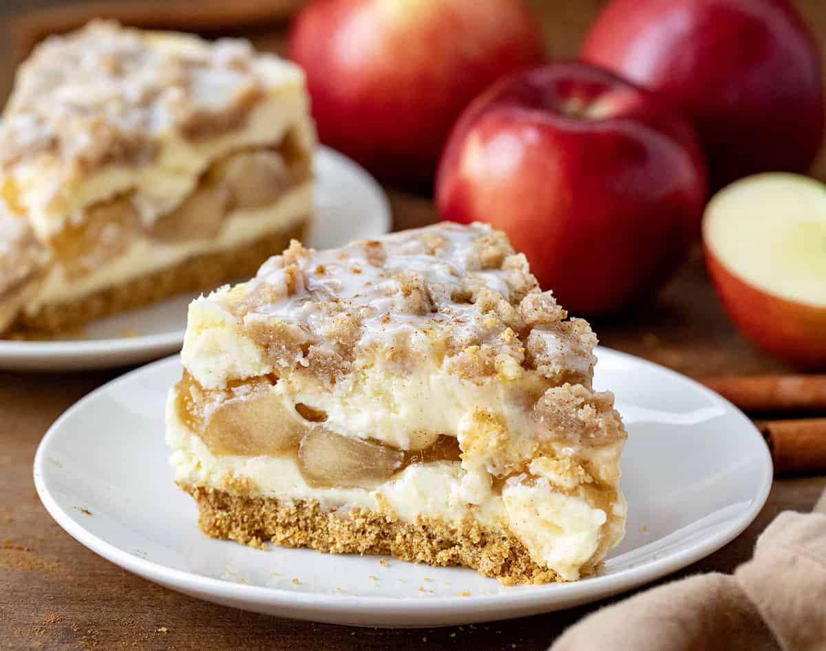 Slice of Apple Crumble Cheesecake on a white plate with another piece in the background with apples.