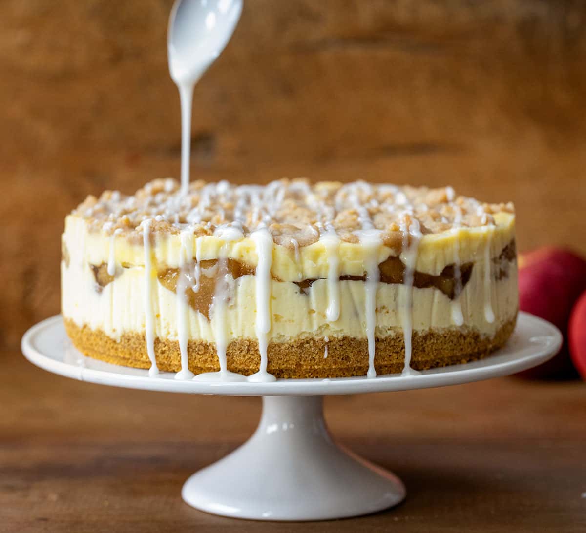 Drizzling glaze over a Apple Crumble Cheesecake on a white cake stand on a wooden table.