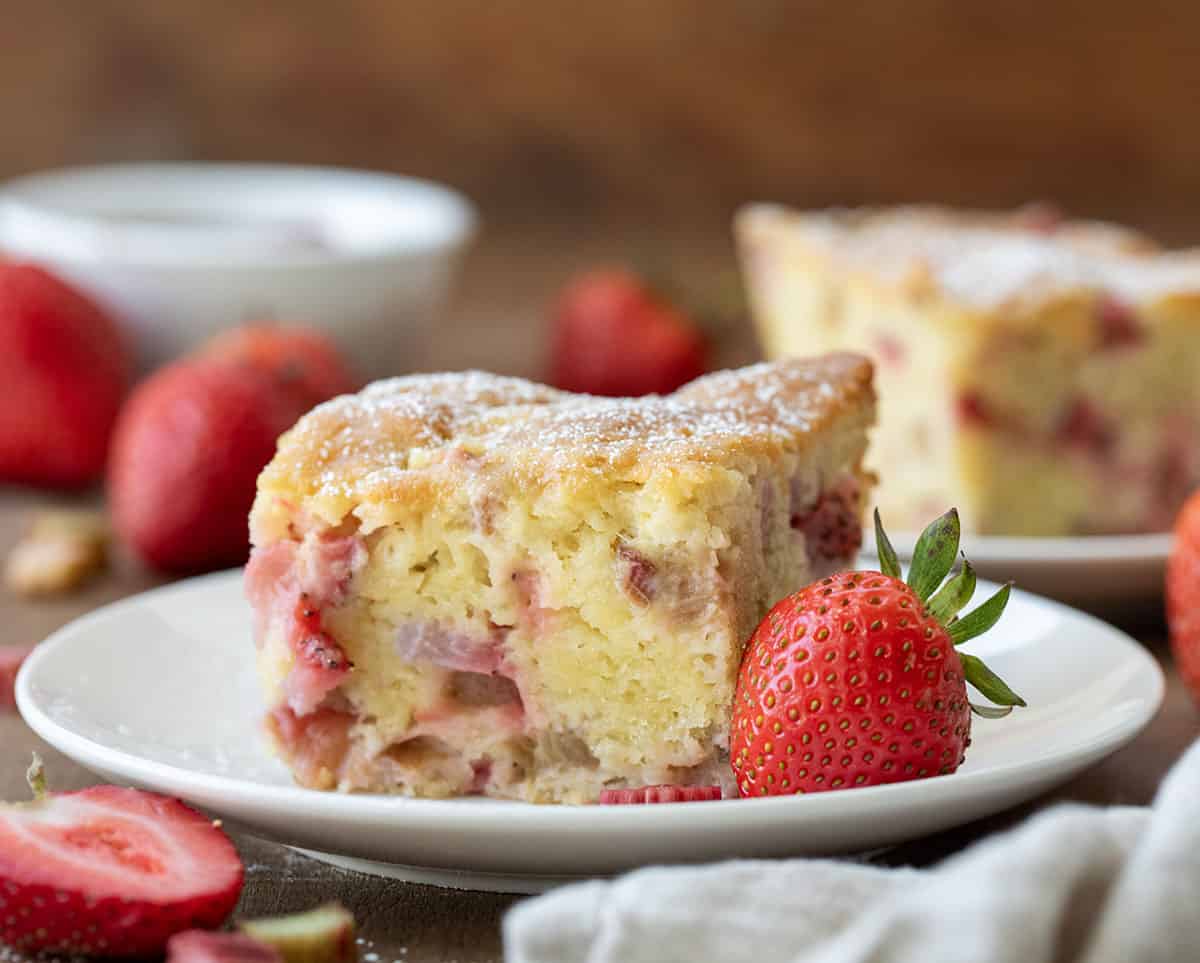 Piece of Fresh Strawberry Rhubarb Cake on a white plate with a fresh whole strawberry.