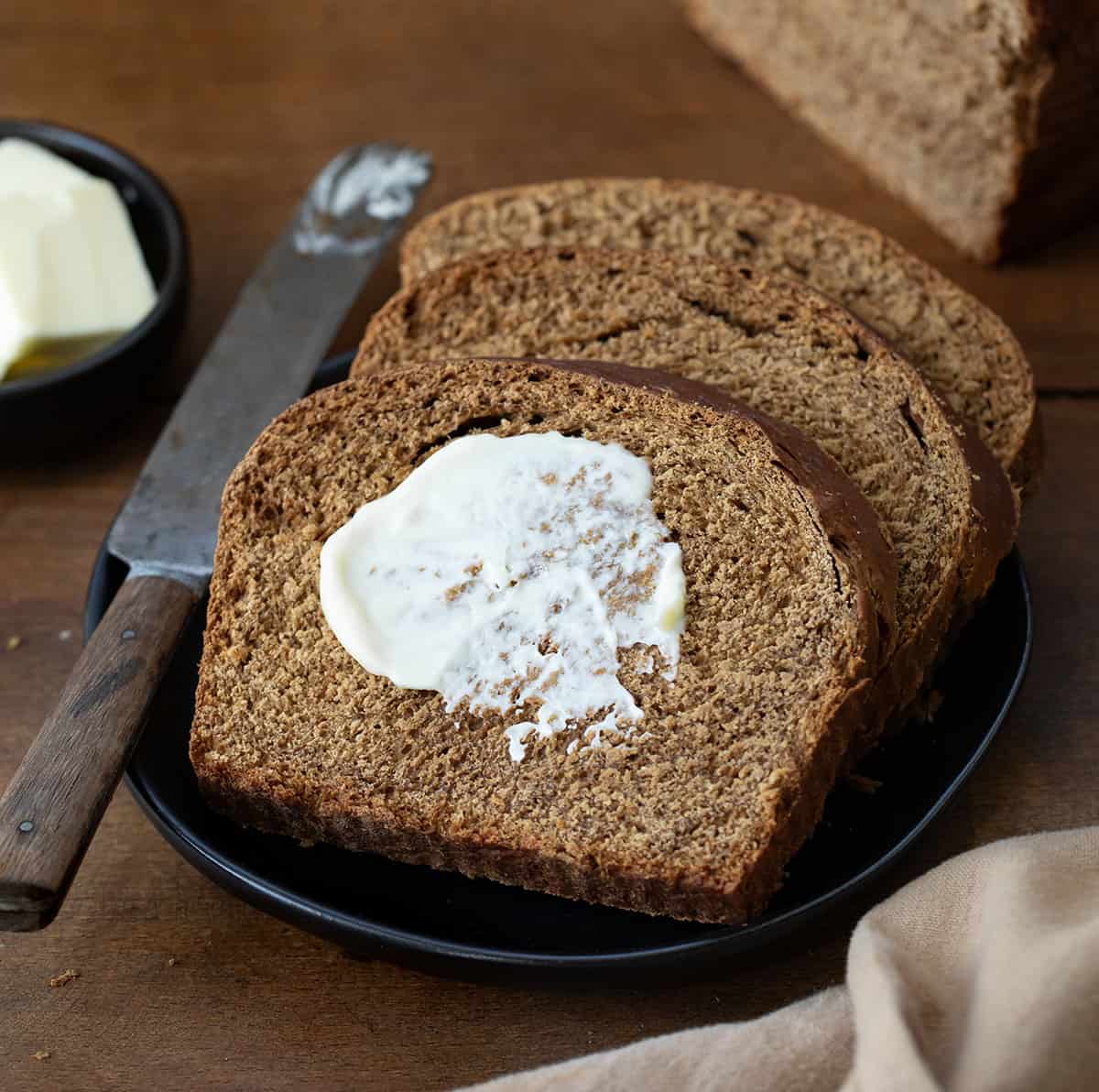 Slices of Pumpernickel Bread on a black plate with butter on the top piece and a knife resting on the plate.