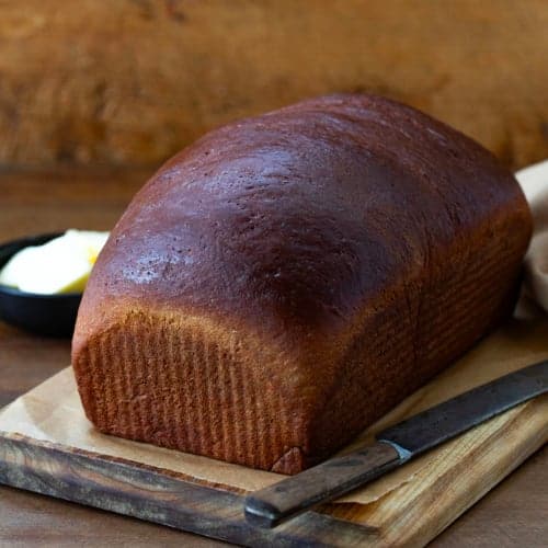 Whole Pumpernickel Bread loaf on a cutting board on a wooden table.